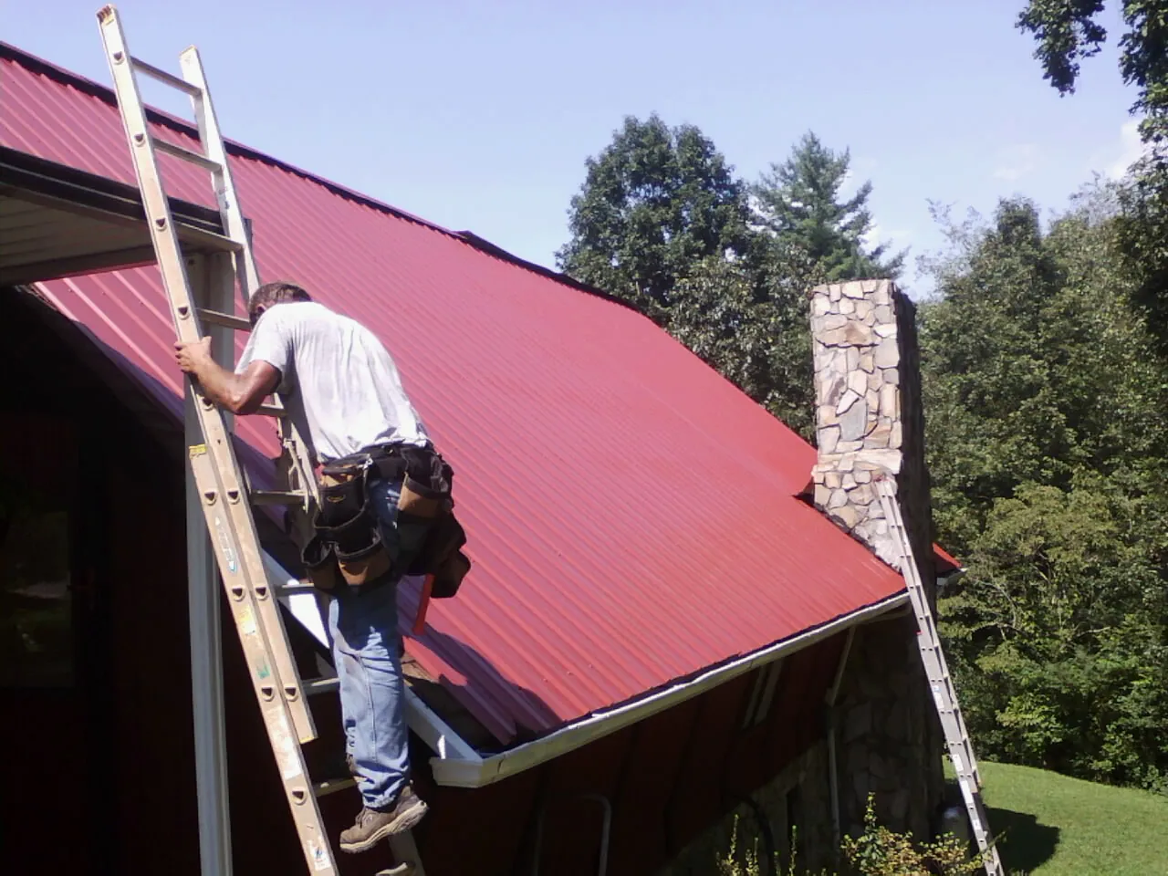 Roofer on a ladder beside a red metal roof and stone chimney — Soffit & Fascia Repair by Roofing Brothers in Chemult